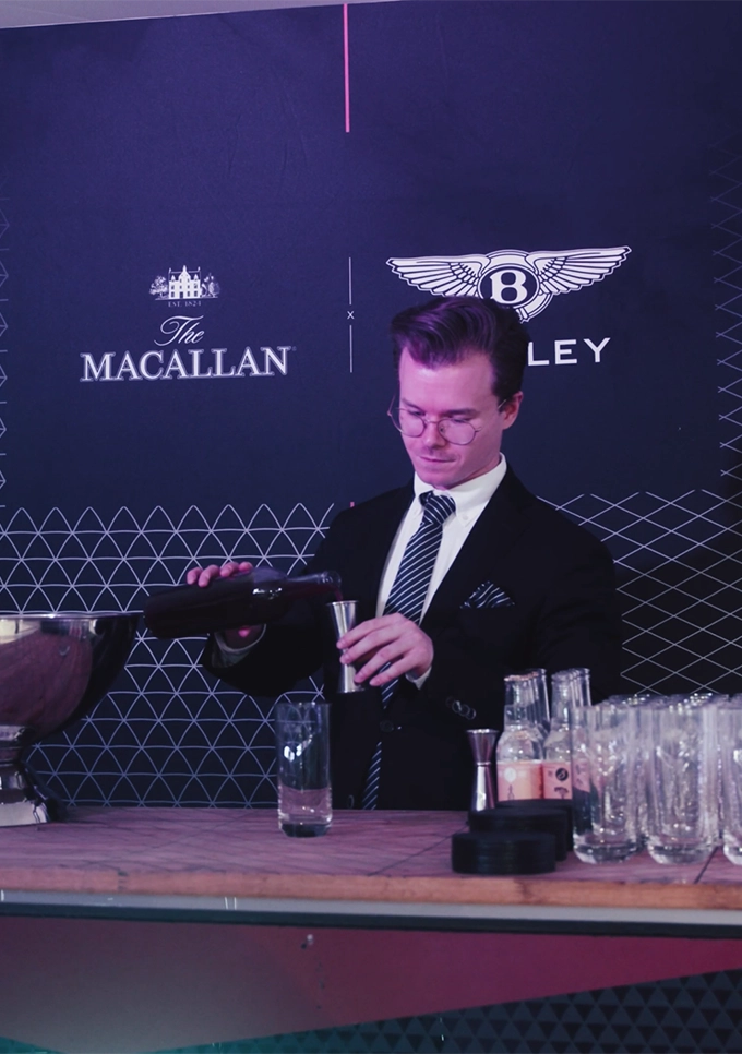 Bartender i kostym blandar drink bakom bar under Bentley-event, med logotyper för Bentley och The Macallan i bakgrunden
