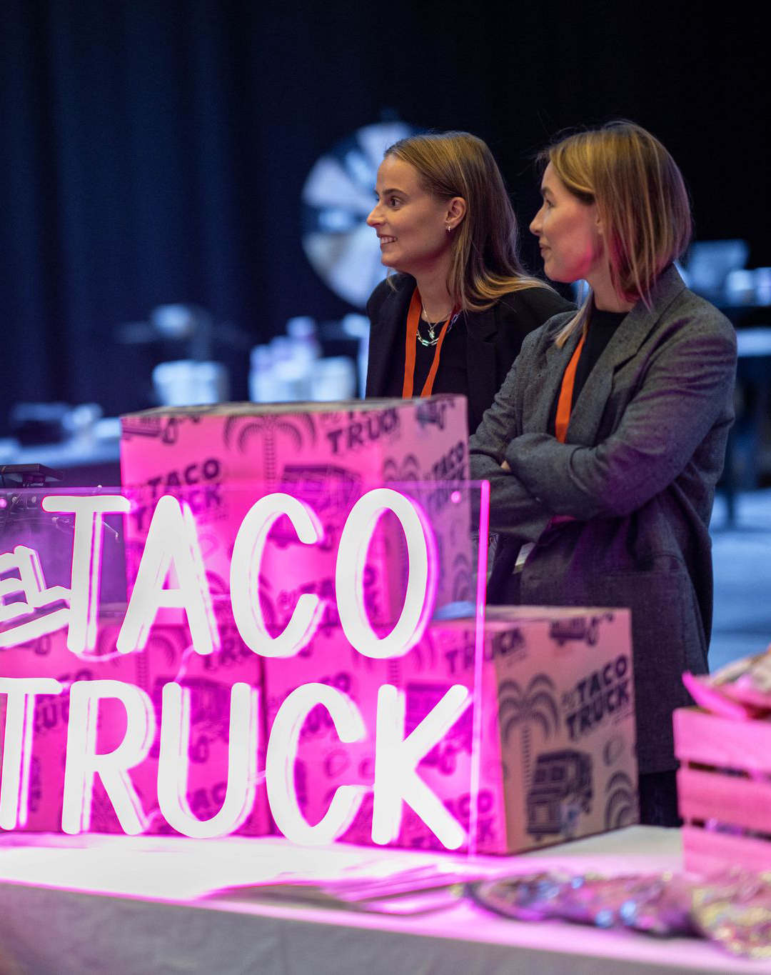 Tre personer står och samtalar vid en färgglad monter från El Taco Truck, upplyst av ett rosa neon-skylt.