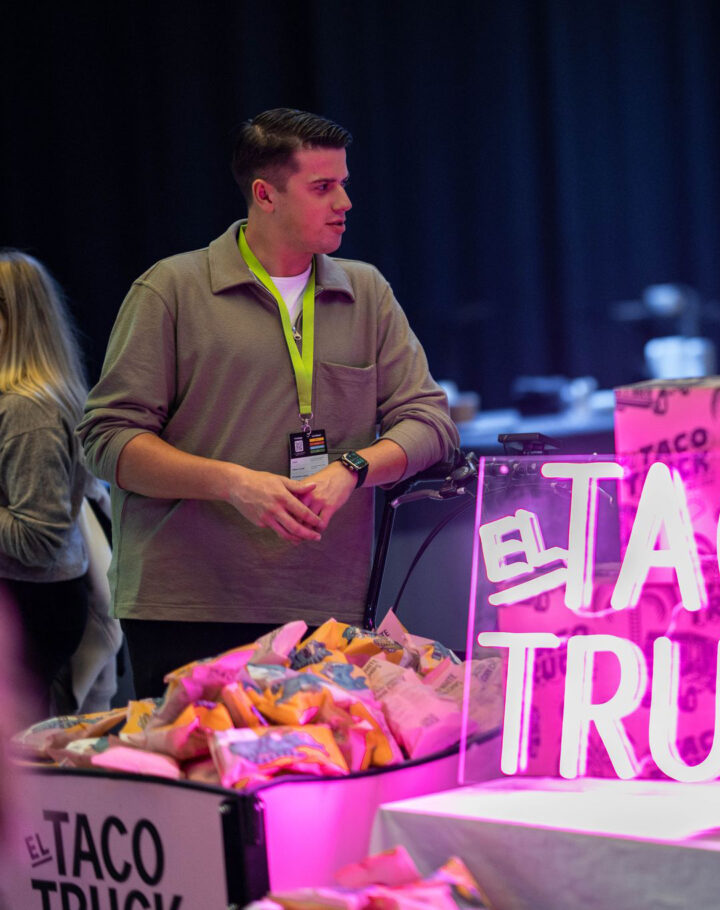 En personer står och samtalar vid en färgglad monter från El Taco Truck, upplyst av ett rosa neon-skylt.
