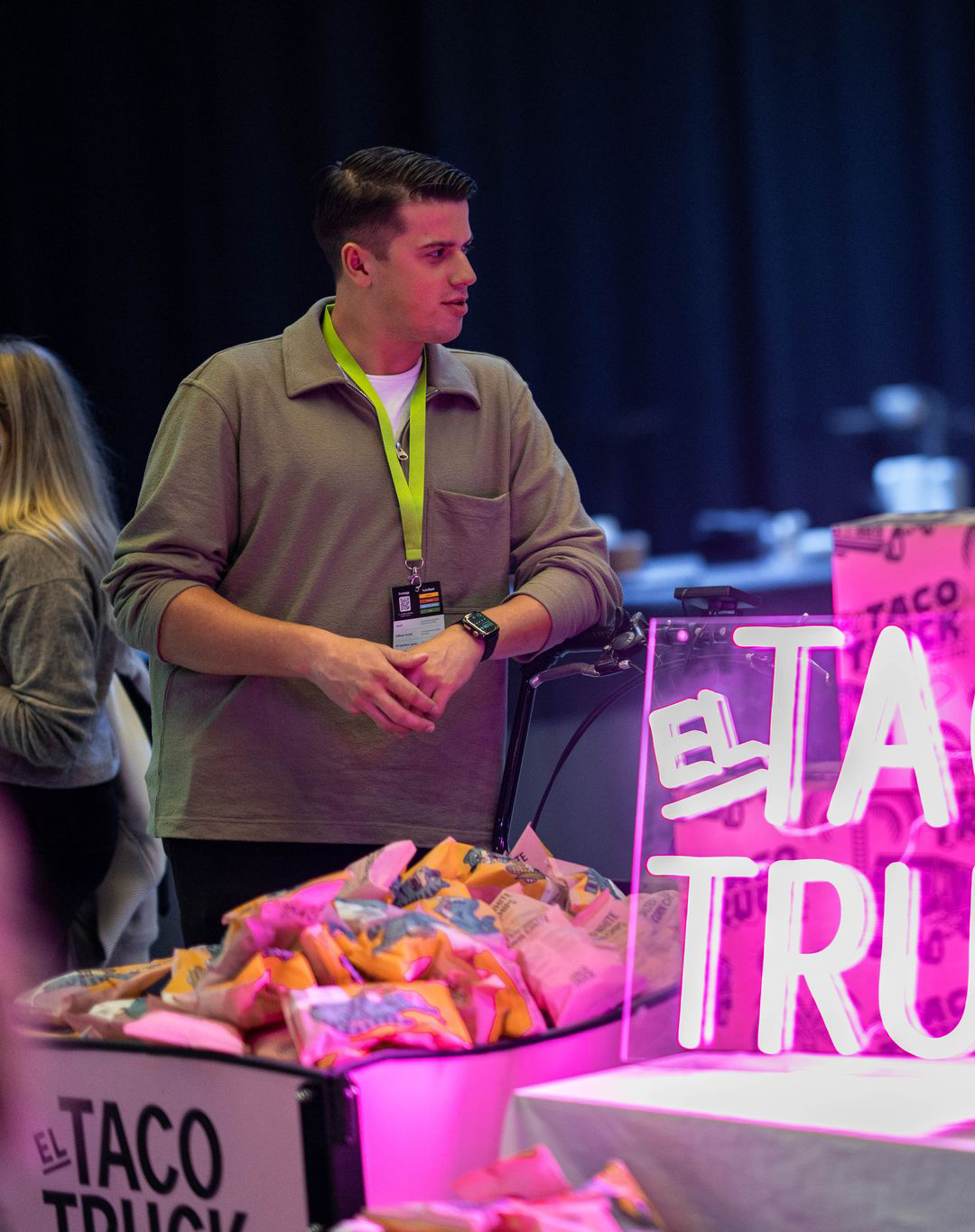 En personer står och samtalar vid en färgglad monter från El Taco Truck, upplyst av ett rosa neon-skylt.