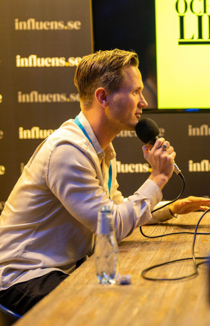 Viktor Frisk sitter vid ett bord och håller i en mikrofon under en podcast- eller intervjuinspelning, med influens.se som bakgrund.