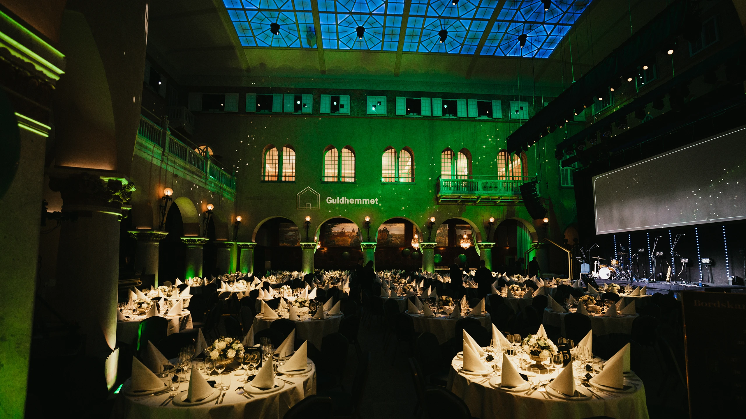 Guldhemmet-galan i Stockholms stadshus med dukade runda bord, scen och grön ljussättning i en pampig eventmiljö