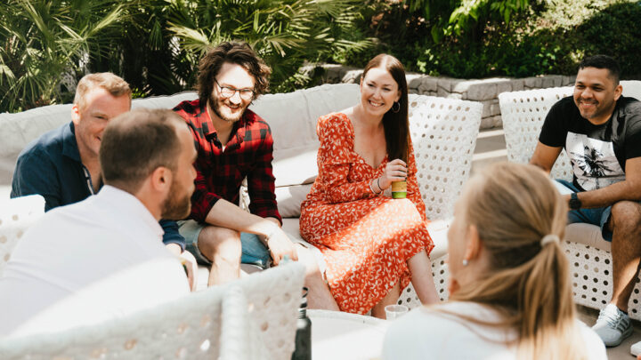 En grupp människor sitter utomhus i en loungegrupp och pratar tillsammans i en solig miljö.