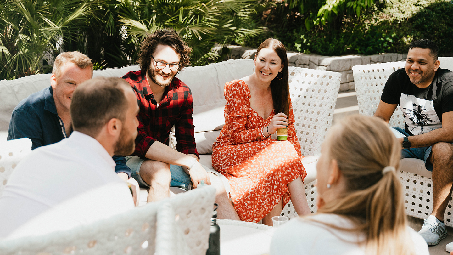 En grupp människor sitter utomhus i en loungegrupp och pratar tillsammans i en solig miljö.