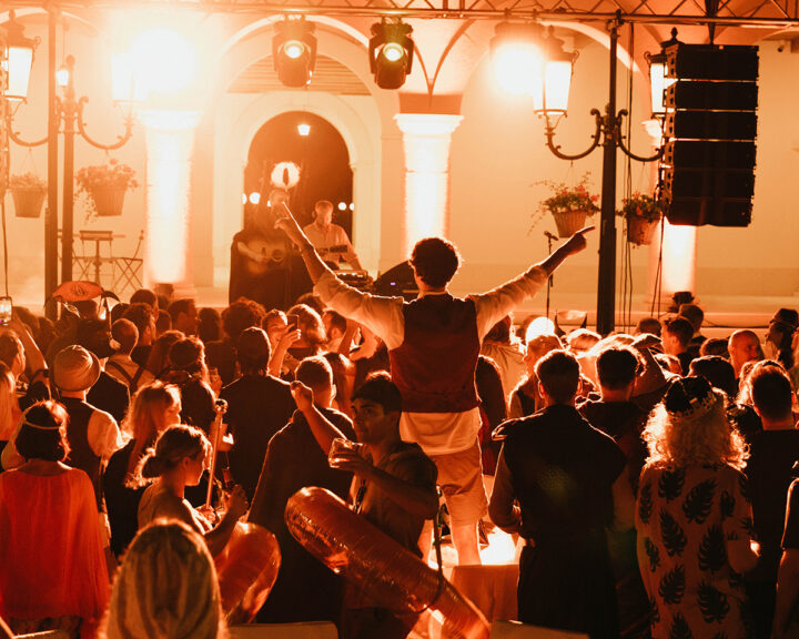 En stor grupp människor festar framför en scen med starkt varm belysning i en utomhusmiljö på kvällen.