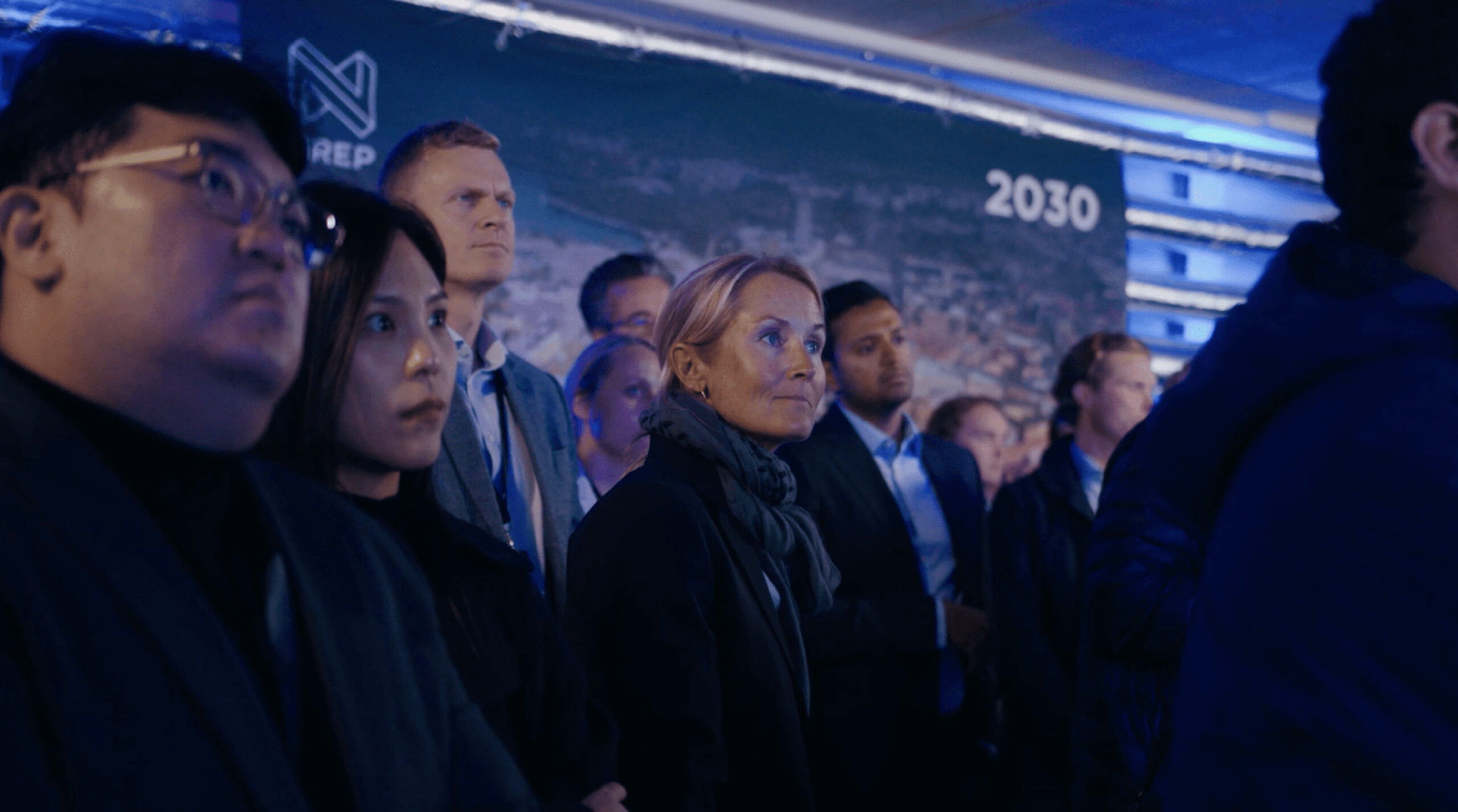 En grupp personer står samlade i en mörkblå belyst miljö och lyssnar uppmärksamt vid ett event.