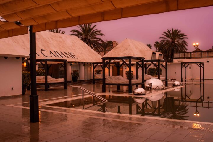 Poolområde i Marbella i skymning, med vita cabanas, spegelblanka vattenytor, palmer i bakgrunden och varma ljus som reflekteras i poolen.