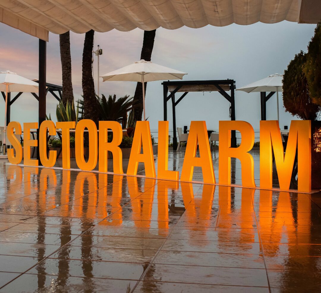 Upplysta bokstäver som stavar Sector Alarm vid en awardgala i Marbella, placerade på en terrass med palmer och havet i bakgrunden i kvällsljus.