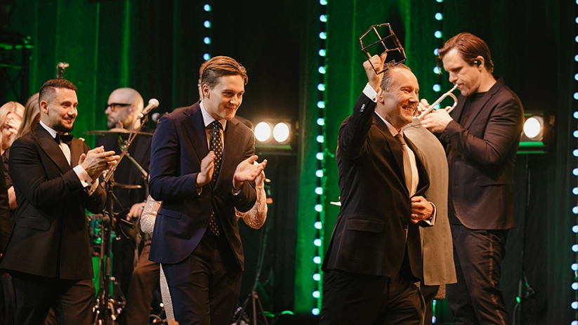 Vinnare på scen under Guldhemmet-galan som håller upp pris och firas med applåder, med liveband och scenljus i bakgrunden