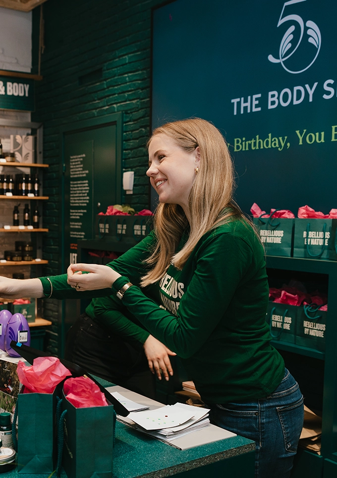 Personal i The Body Shop-butik delar ut produkt till kund under 50-årsjubileum, med brandade goodiebags och kampanjbudskap i bakgrunden.