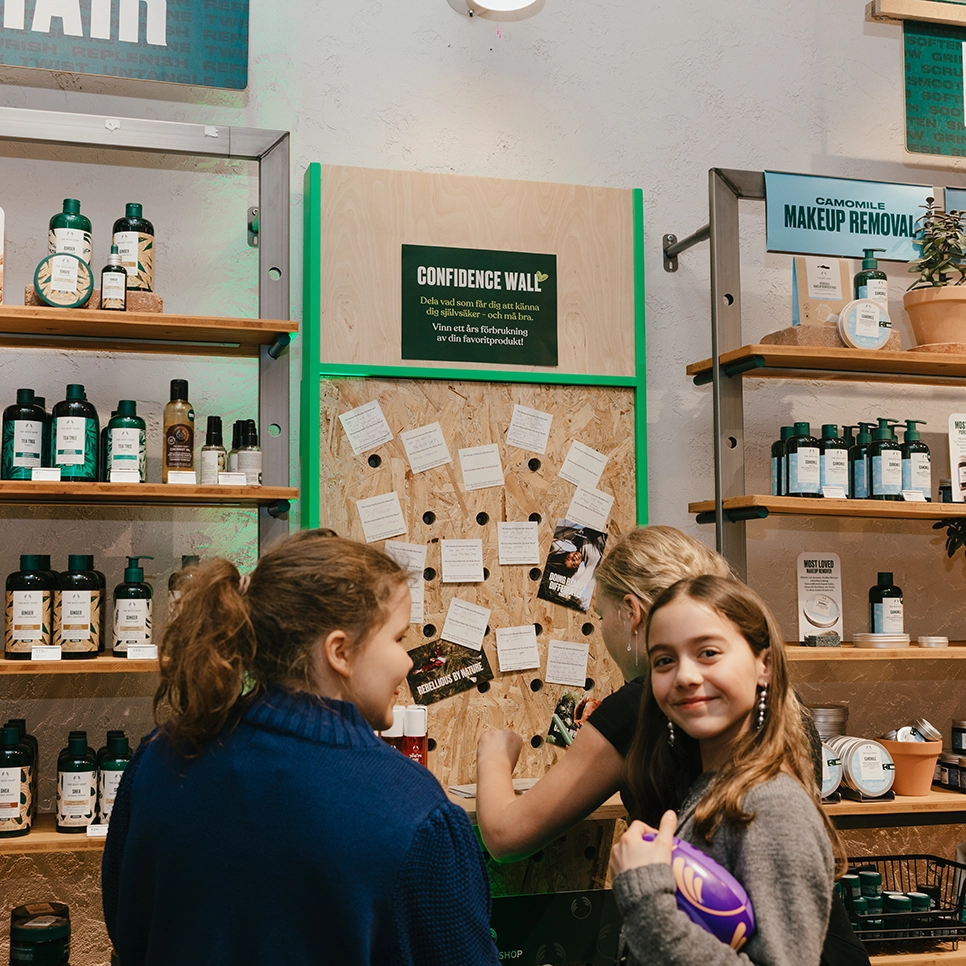 Besökare skriver och sätter upp lappar på “Confidence Wall” i The Body Shop-butik under 50-årsjubileum, omgivna av hudvårdsprodukter och kampanjmiljö.