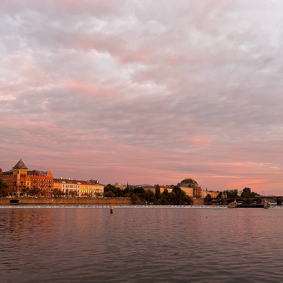 Kvadrat i Prag. Prags byggnader längs floden Vltava i varmt kvällsljus under en rosafärgad himmel.